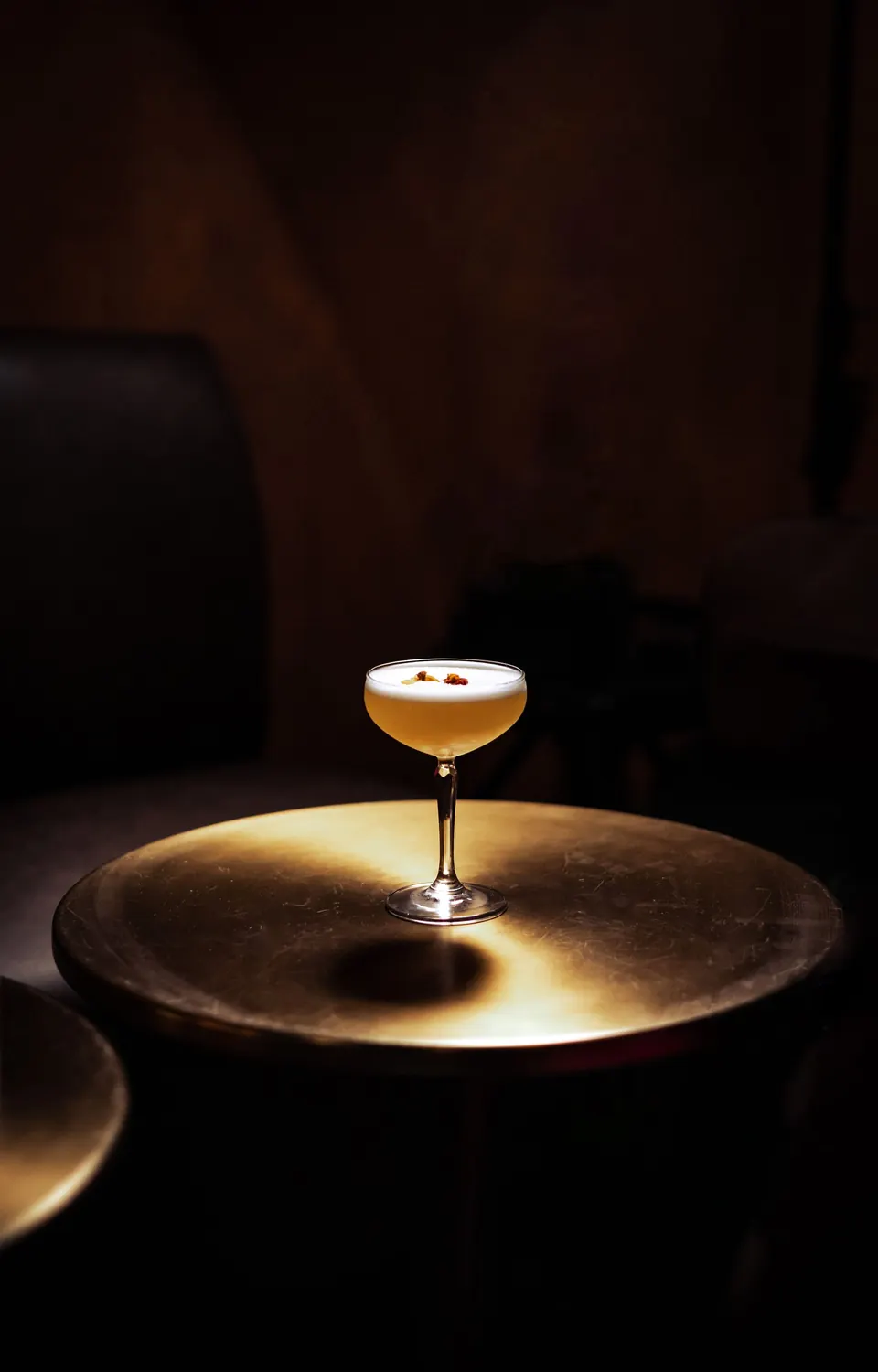 A single foamed cocktail in a coupe glass on a dim, polished table