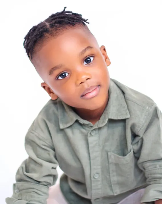 Young boy with short twists in a sage-green corduroy shirt, leaning towards the camera against a white backdrop