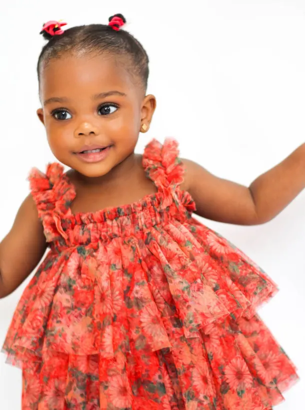Smiling toddler in a tiered red floral tulle dress, hair in two small puffs, against a white studio backdrop
